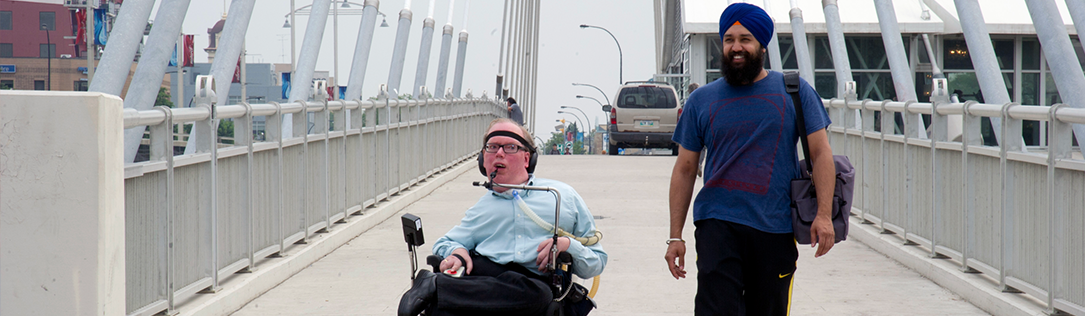 A person sits in a wheelchair and another person stands to his left on a bridge.