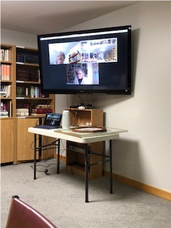 A TV attached to the wall within a meeting space. On the TV their is a Zoom meeting in progress. 