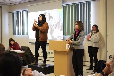 A group of people in attendance at one of the mental health workshops. A women is standing at a podium and another one is providing ASL interpretation of the presentation. 