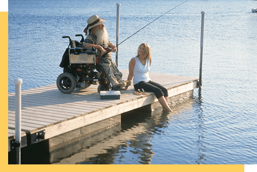 Une personne en fauteuil roulant pêche sur un quai en compagnie d‘une personne assise sur le quai.