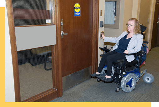 Une personne en fauteuil roulant appuie sur le bouton d‘accès d‘une porte.