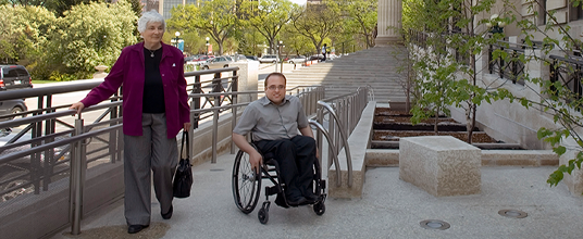 A person in a wheelchair and a person walking on a sidewalk.